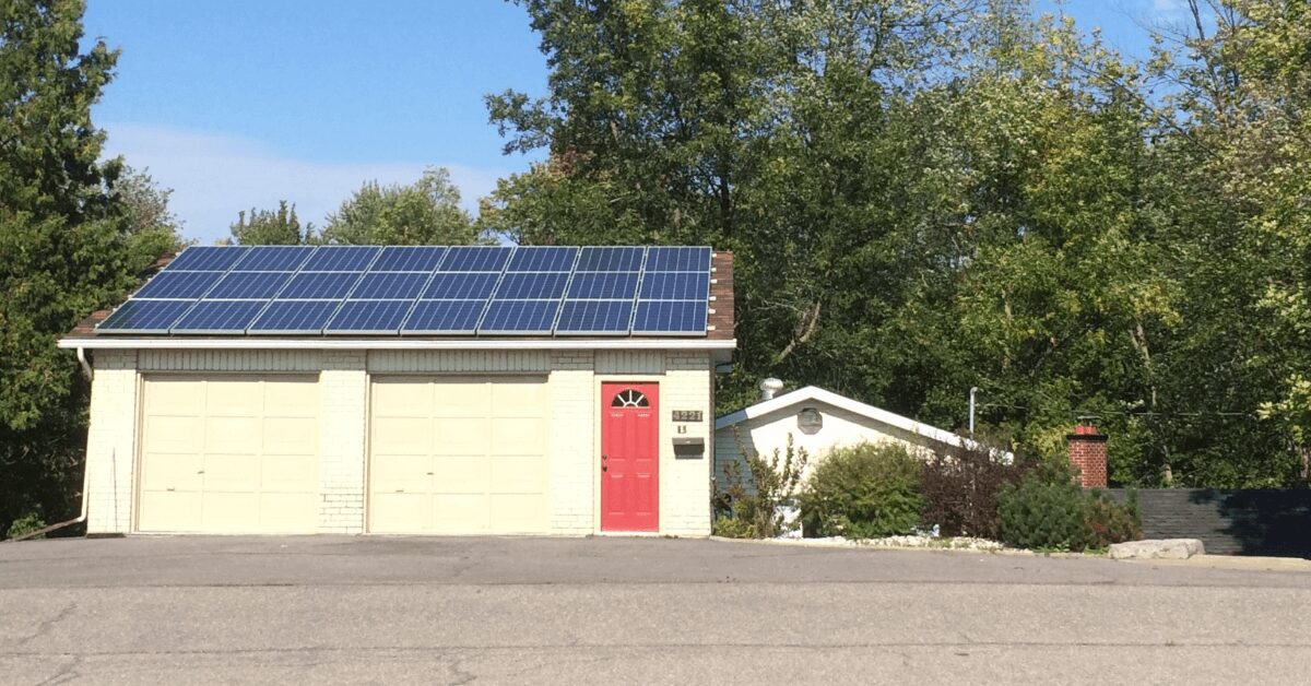 Garage-style building with three cream doors, a bright red front door, and solar panels on the roof; a small white shed sits to the right with trees behind.