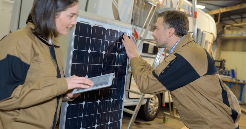 Two technicians in beige jackets examine a solar panel in a workshop, one holding a tablet nearby.