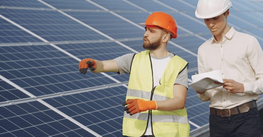 Two solar farm workers in safety gear inspect a photovoltaic panel array; one points as the other reviews plans.