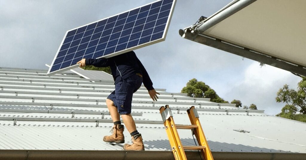 Worker on a ladder lifts a solar panel onto a metal roof for installation