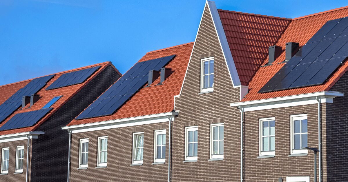 Row of brick townhouses with red tile roofs and solar panels under a blue sky in a sunny neighborhood.