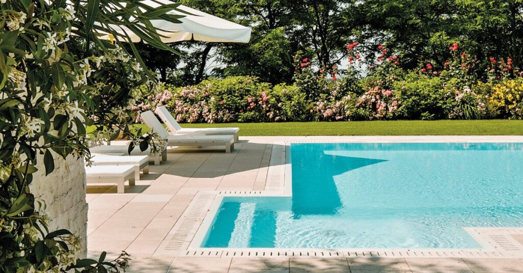 Outdoor poolside scene with turquoise water, white loungers under an umbrella, and a garden of blooming flowers in the background.