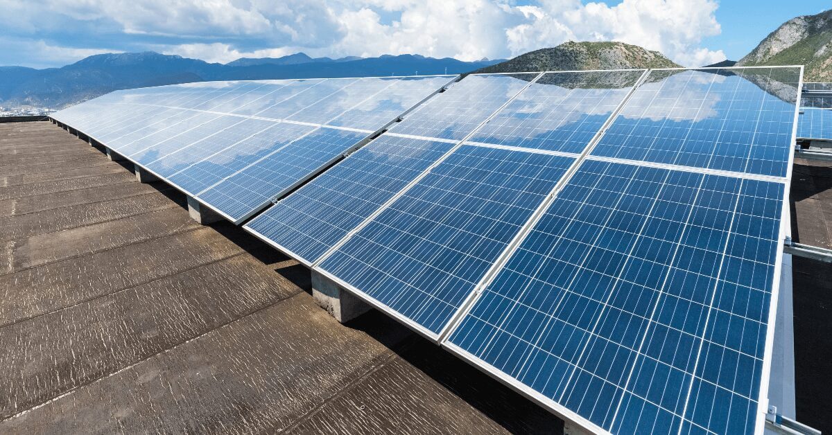 Rows of blue solar panels mounted on a sloped rooftop with mountains in the distance under a partly cloudy sky, sunlight reflecting off the panels.