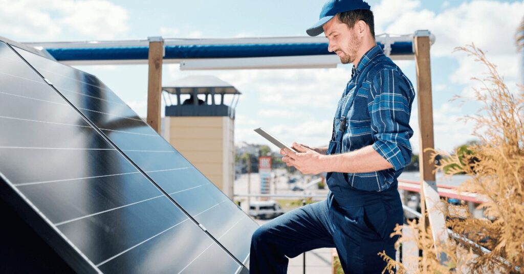 Worker in a blue plaid shirt and cap holds a tablet while inspecting solar panels outdoors at a sunny site.