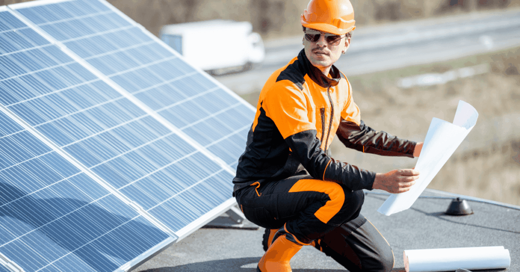 Worker in orange safety gear and hard hat examining blueprints on a rooftop beside solar panels, wearing safety glasses.