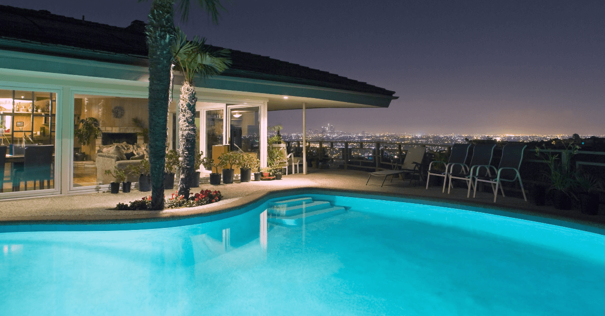 Night view of a lit backyard pool beside a modern house, with a city skyline in the distance and patio loungers on the deck.
