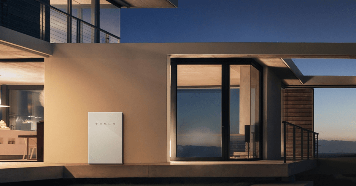 Modern house exterior at dusk with a Tesla Powerwall mounted on the wall beside glass doors and large windows behind.