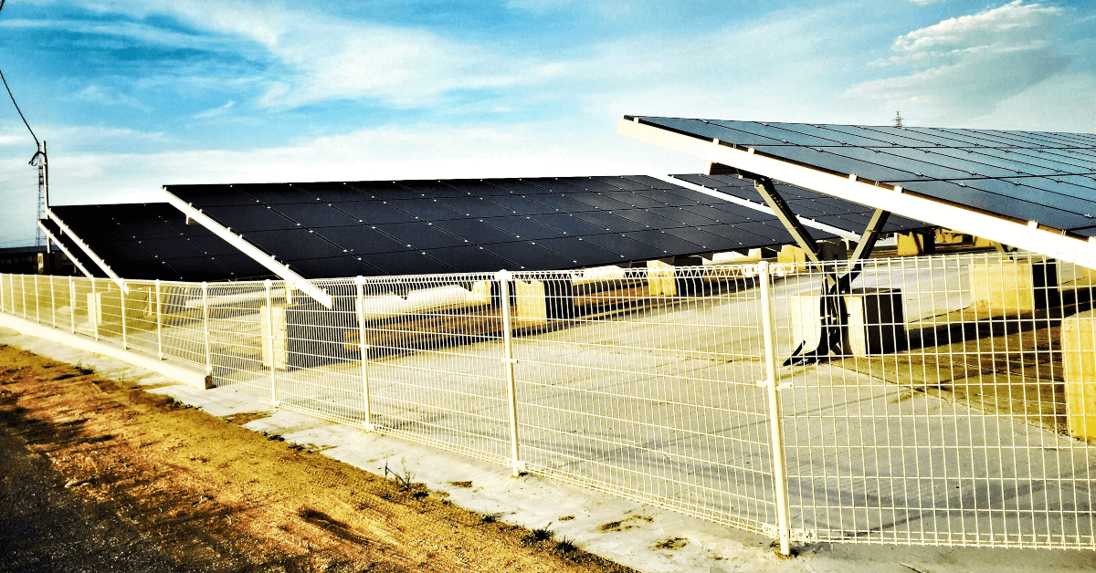 Row of solar panels tilted on a rooftop behind a white fence under a blue sky.