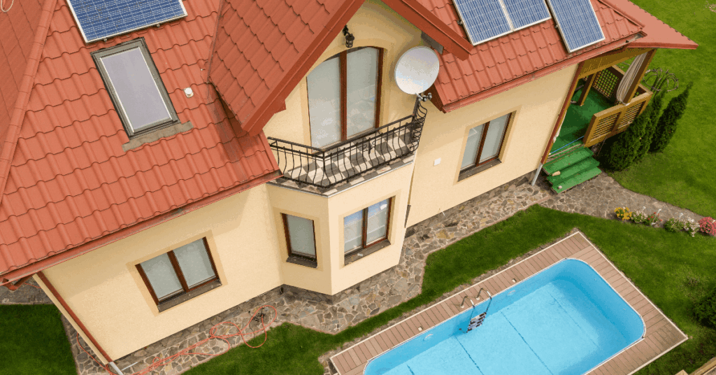 Aerial view of a beige, two‑story house with a red tile roof, solar panels, and a small balcony, with a blue rectangular pool in the yard.