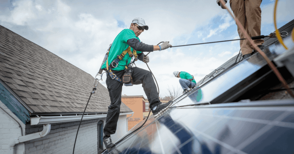 Two workers in green shirts with harnesses roof-cleaning or repairing a sloped roof, secured by ropes against a blue sky signifying safety-first roof work