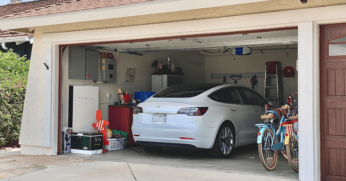 White Tesla sedan parked in an open garage with bicycles on the right and cluttered storage to the left/far wall.