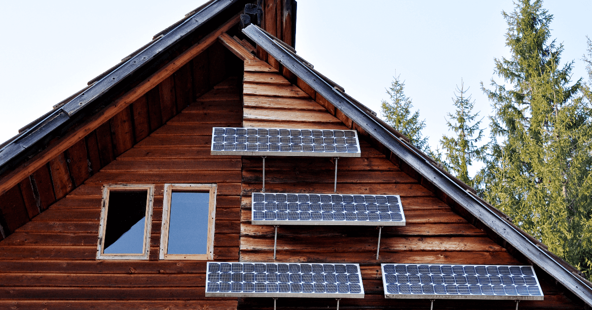 Wooden cabin with three rows of solar panels mounted on its exterior wall, against a blue sky and pine trees.