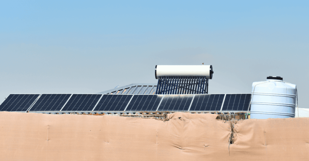 Row of solar panels on a roof with a solar water heater behind them and a large blue water storage tank on the right under a clear blue sky.