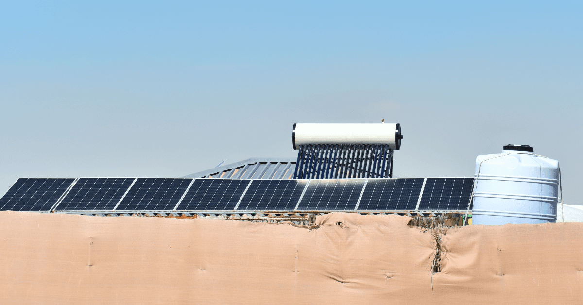 Row of solar panels on a roof with a solar water heater behind them and a large blue water storage tank on the right under a clear blue sky.