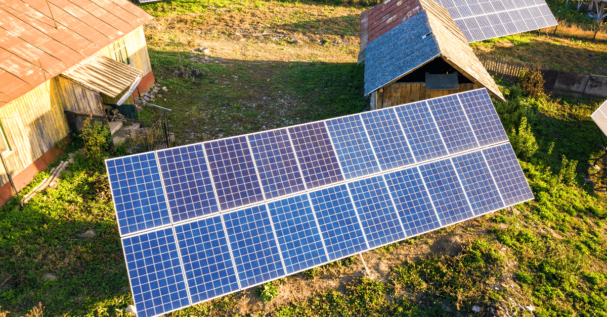 Solar panel array installed on a grassy yard beside rural buildings (aerial view).