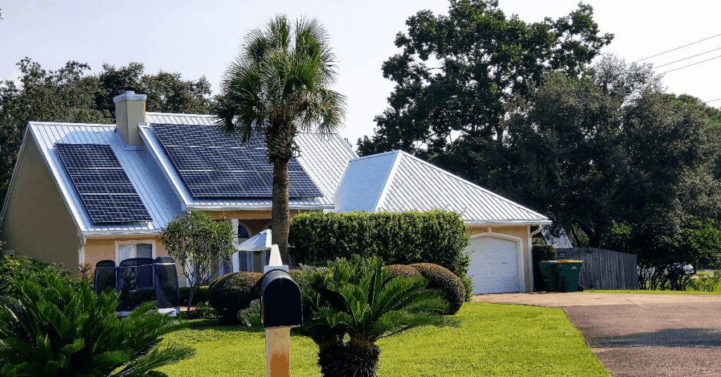 Residential house with solar panels on a metal roof, palm tree in front yard, and a driveway with a garage door.