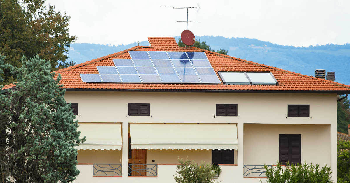 Residential house with solar panels on a red-tiled roof and a satellite dish in a mountainous backdrop.
