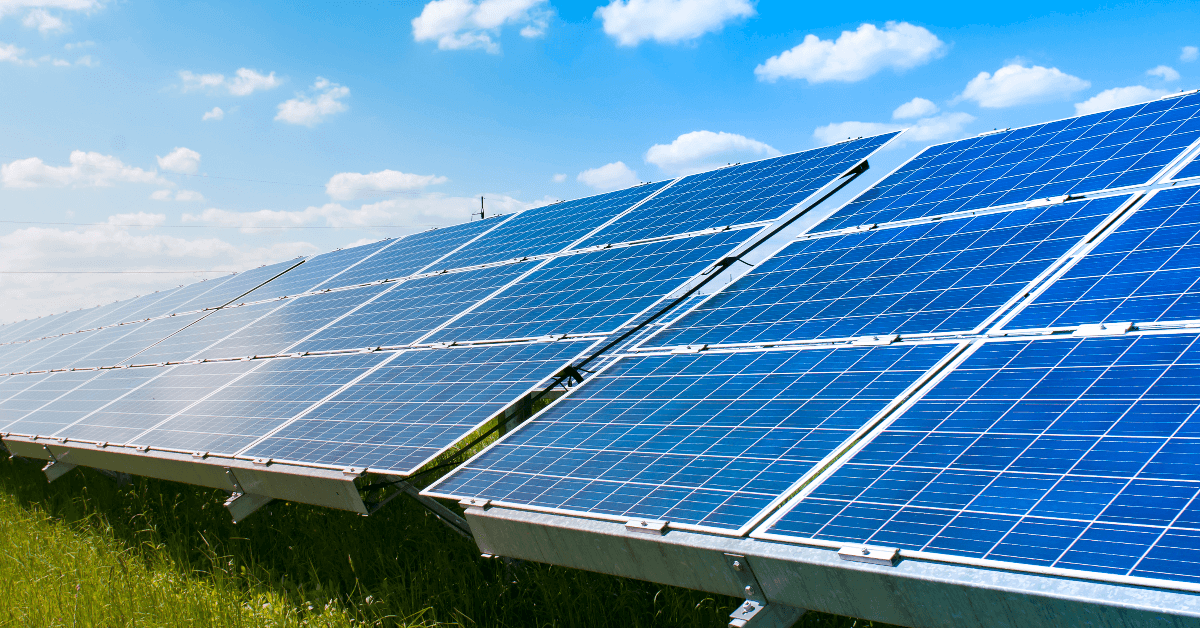 Rows of blue solar panels in a grassy field under a bright blue sky with scattered clouds, representing solar energy installation.