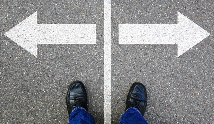 Person wearing black shoes stands on pavement at a fork with large white left and right arrows diverging.