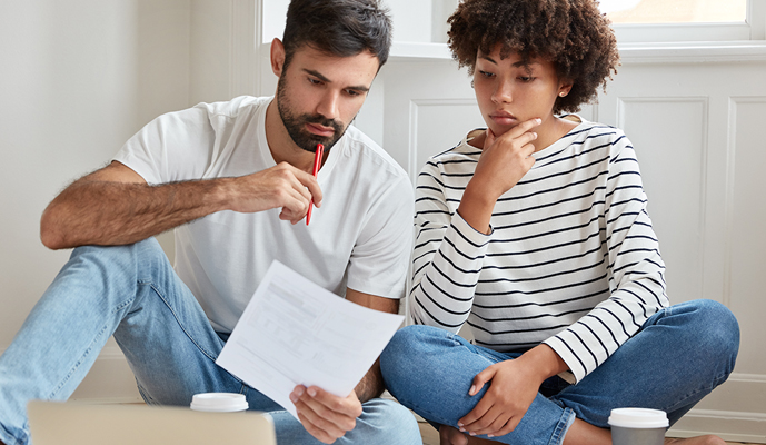 Two adults sit on a floor, reviewing a document together with a red pen in hand and coffee cups nearby.