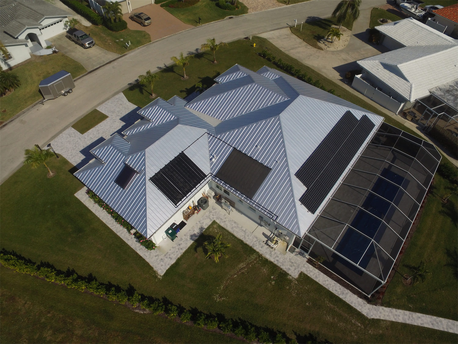 Aerial view of a modern suburban house with a multi-section gray metal roof and a large solar panel array on the screened patio.