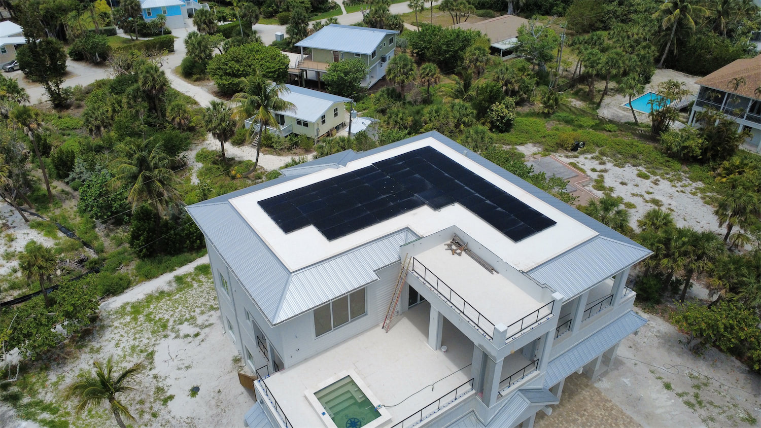 Aerial view of a large white house with black solar panels on its flat roof, surrounded by palm trees and a tropical neighborhood.