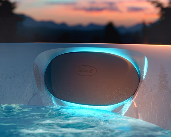 White oval device mounted at a pool edge, glowing blue LEDs with a sunset and mountains in the background.