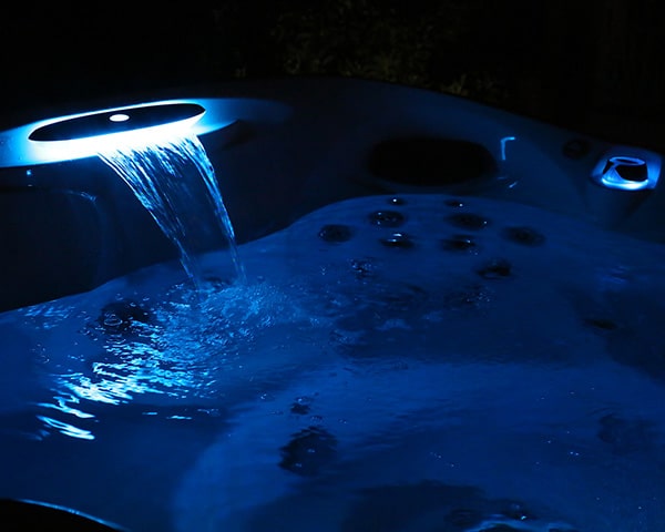 Blue-lit hot tub with water cascading from a spillover edge, jet bubbles visible and night scene setting.