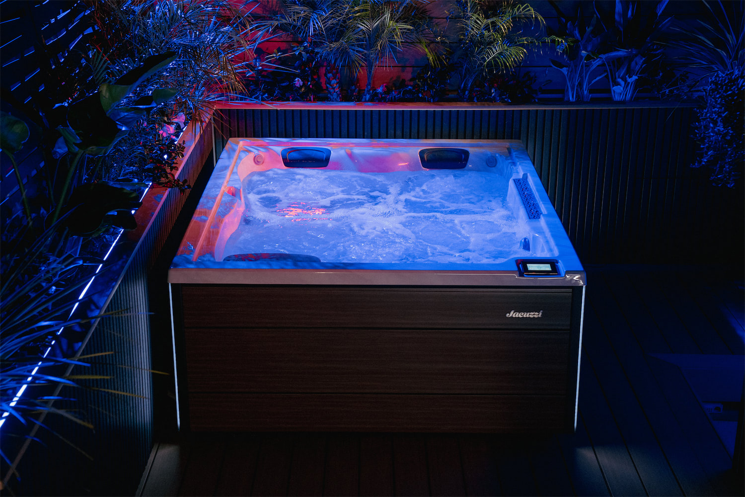 Outdoor hot tub on a wooden deck at night, illuminated by blue lighting with surrounding tropical plants and a privacy fence nearby.