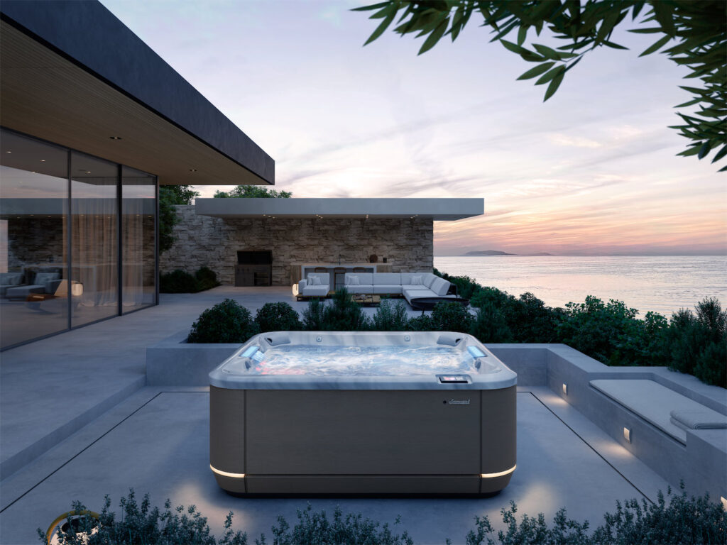 Modern outdoor terrace by the sea with a lit hot tub in the foreground and a stone wall lounge area at sunset.
