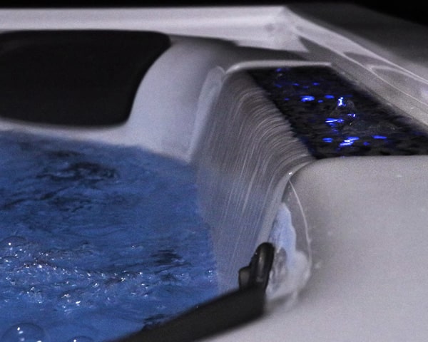 Close-up of a white hot tub edge with water cascading over a spillway and blue LED lighting.