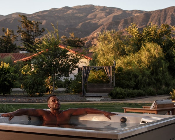 Man relaxing in an outdoor hot tub at sunset, with trees, a wooden gazebo, and distant mountains in the background.