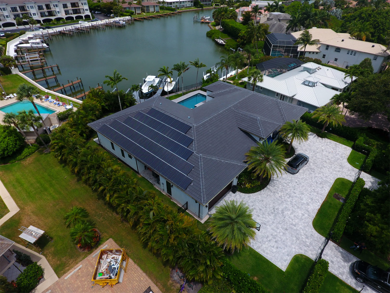 Aerial view of a modern house with a solar-panel roof, palm trees, and a canal with boats nearby.