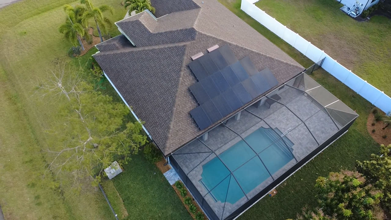Aerial view of a brown-shingled house with solar panels on the roof and a rectangular screened-in pool in the backyard, surrounded by green lawn and a white fence.