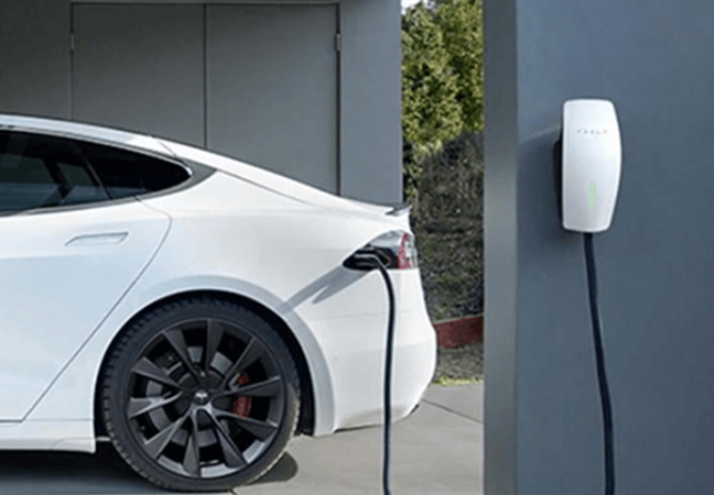 White electric car plugged in to a wall-mounted charging station; charging cable connected at the rear side of the vehicle.