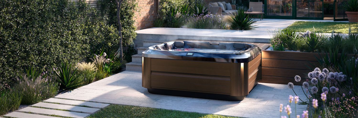 Modern wooden hot tub in an outdoor garden on a stone patio with greenery and flowering plants nearby.