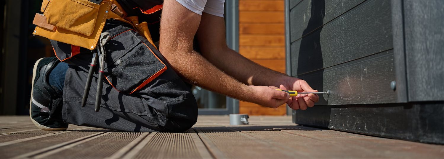 Handyman kneeling on a wooden deck, tightening a screw into dark exterior siding with a screwdriver; tool belt visible.