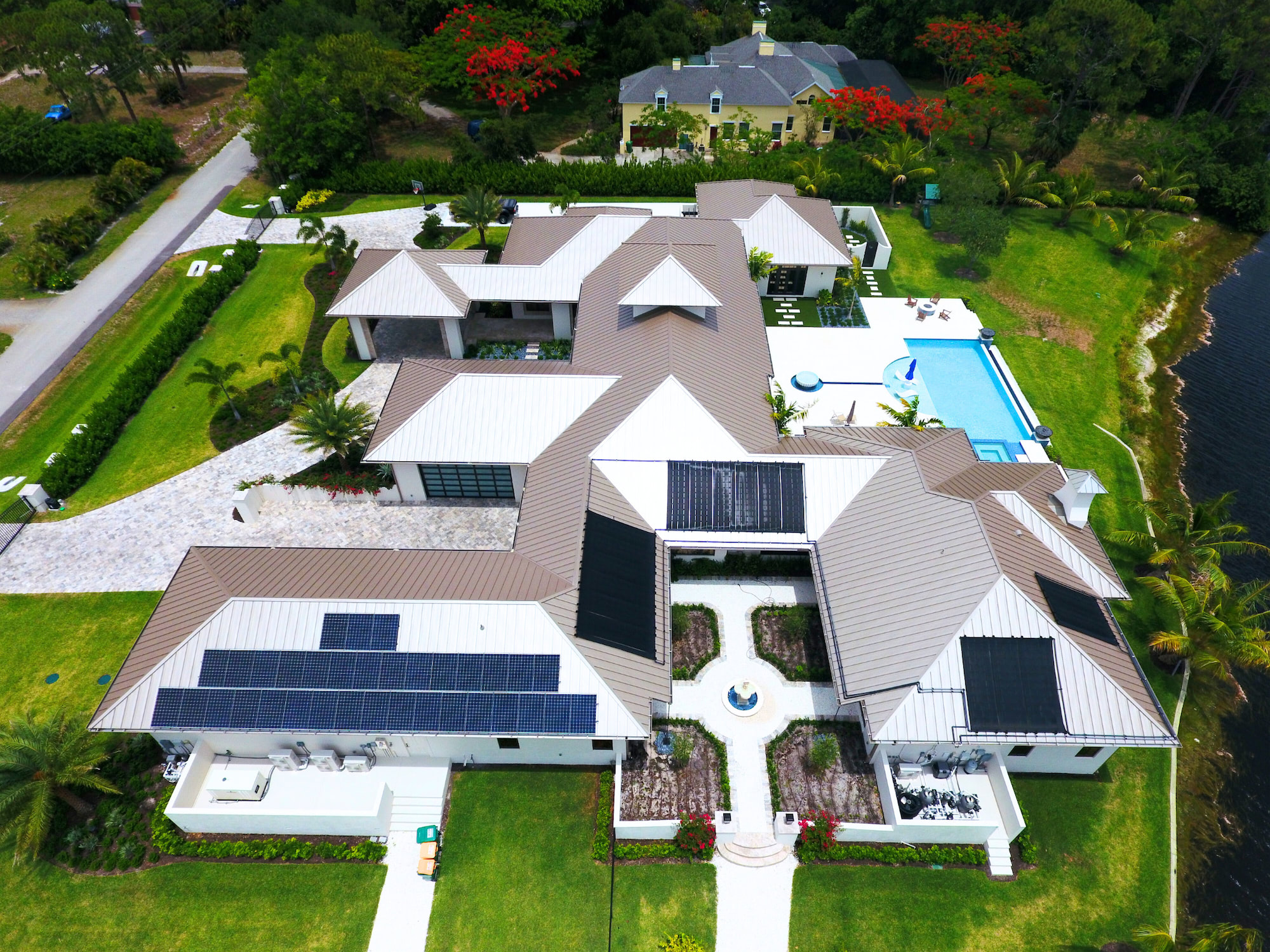 Aerial view of a modern house with multiple white and brown metal roofs, solar panels, a blue rectangular pool, and manicured lawns near a waterfront.