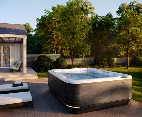 Backyard scene with a square hot tub on a paved patio, lounge chairs, a covered seating area, and a grassy yard with trees in the background.