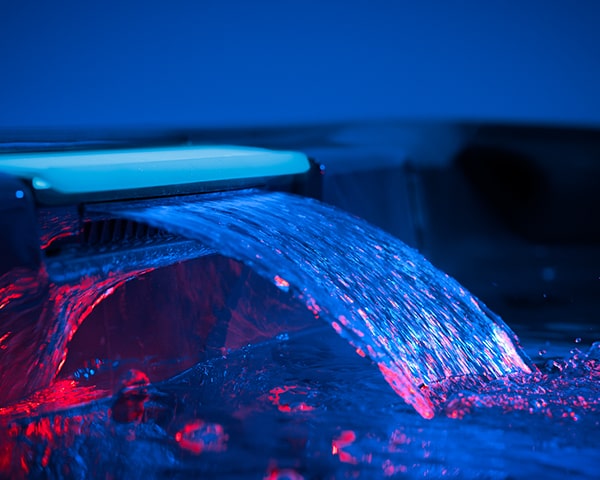 Close-up of a blue-lit water fountain jet arcing into a dark pool, with red reflections on the surface.