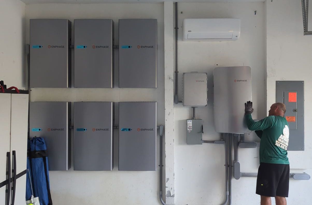 Wall of ENPHASE solar inverters in a utility room; a technician in a green shirt adjusts a large electrical cabinet nearby.