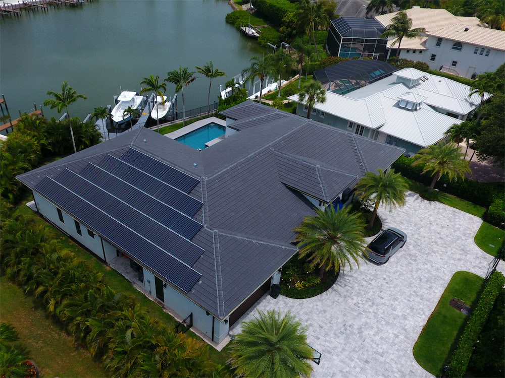 Aerial view of a modern house with solar panels on the roof, a paved driveway, palm trees, and a waterfront canal with boats nearby.