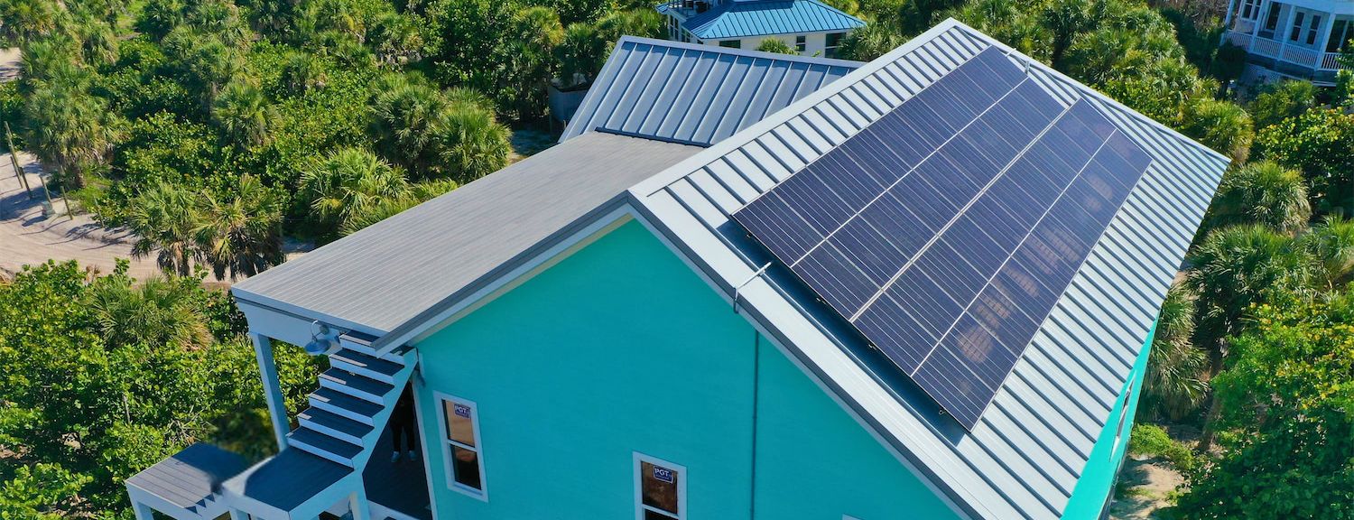Teal house with solar panels on its metal roof, surrounded by palm trees and greenery.