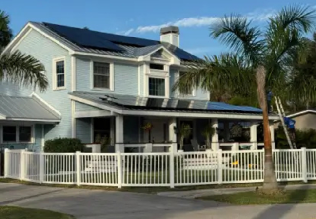 Two-story light-blue house with white trim, solar panels on the roof, a white picket fence, and palm trees in the yard.