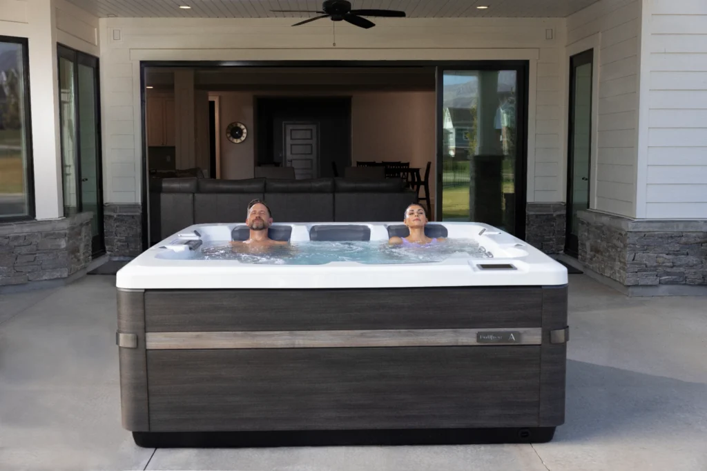 Couple relaxing in a white outdoor hot tub on a covered patio with sliding glass doors to the house interior in the background.